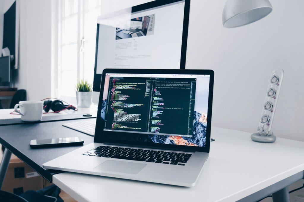 Laptop mit Code auf dem Bildschirm auf einem Schreibtisch, im Hintergrund ein weiterer Monitor, Kaffeetasse und Smartphone, Internetagentur Düsseldorf