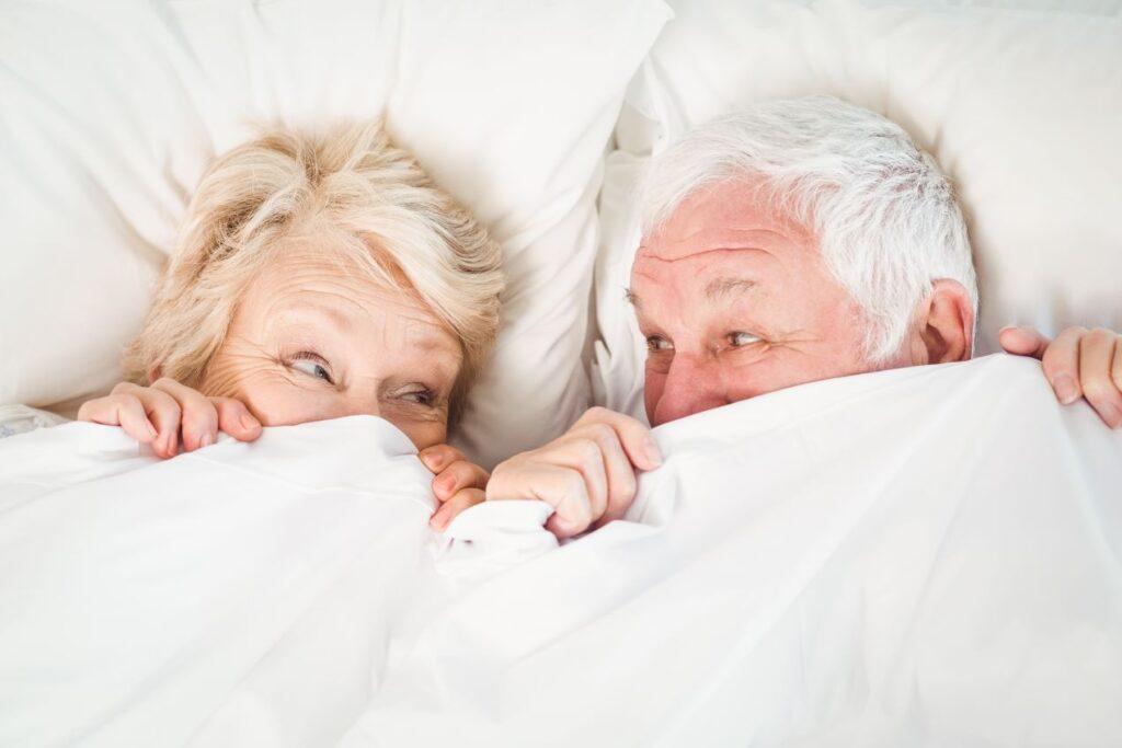 Ein älteres Paar liegt lächelnd unter einer Decke im Bett, das mit einer Pflegematratze ausgestattet ist, und genießt den gemeinsamen Moment