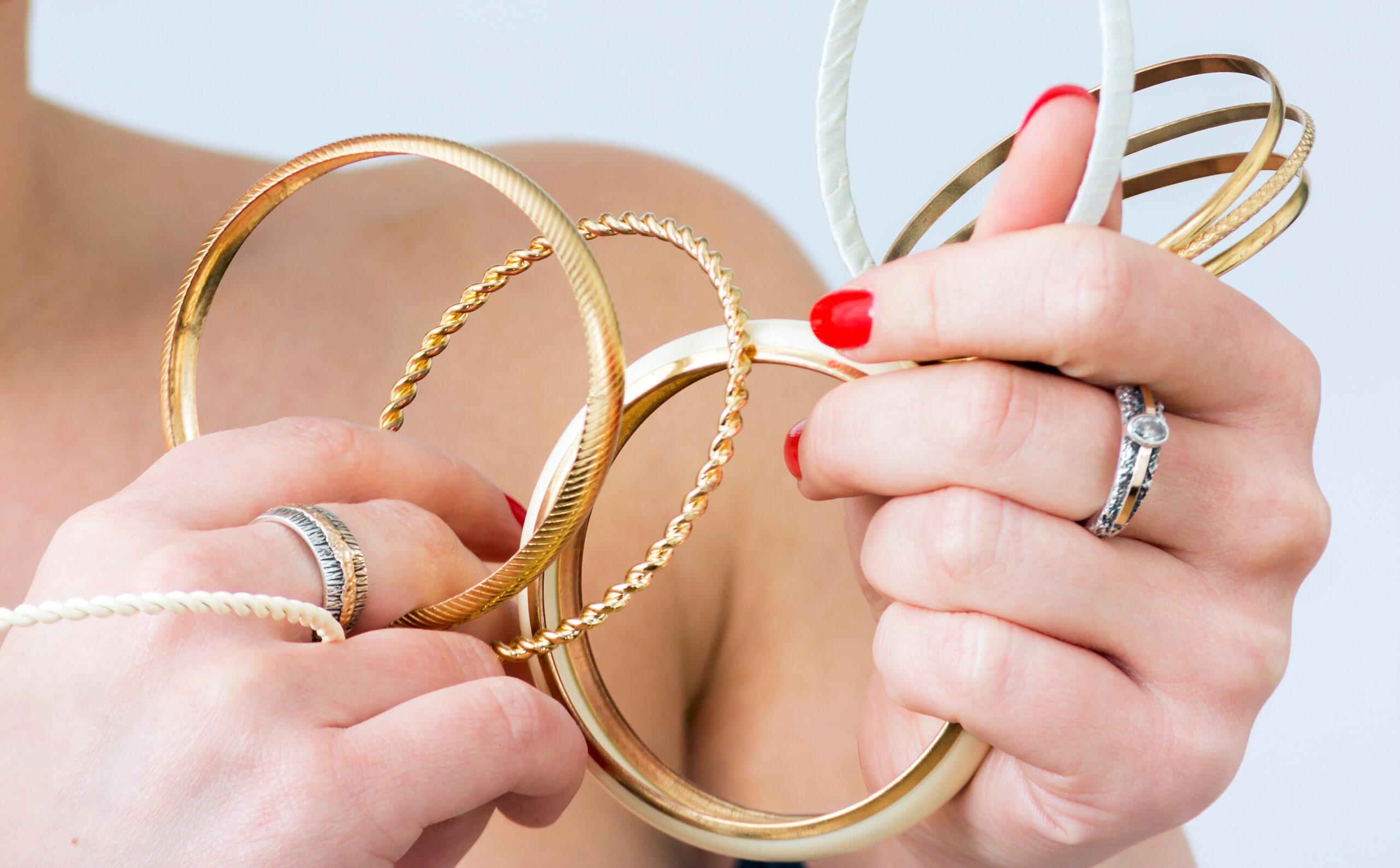 Frau mit roten Nägeln hält eine Auswahl an goldenen Armbändern, darunter schlichte und gedrehte Designs, kombiniert mit silbernen Ringen.